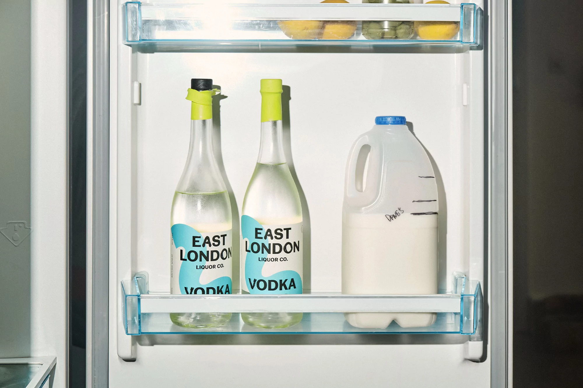 Refrigerator shelf with two bottles of East London Liquor Co. vodka and a carton of milk in a brightly lit indoor setting.