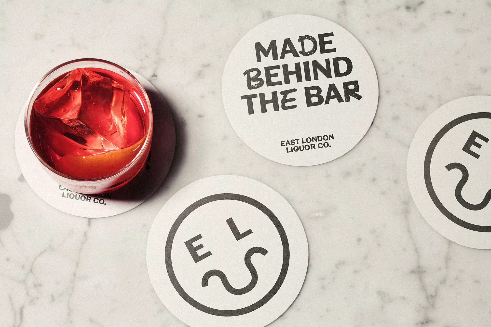 Overhead shot of a red cocktail with ice cubes on a white marble surface, surrounded by East London Liquor Company branded coasters.