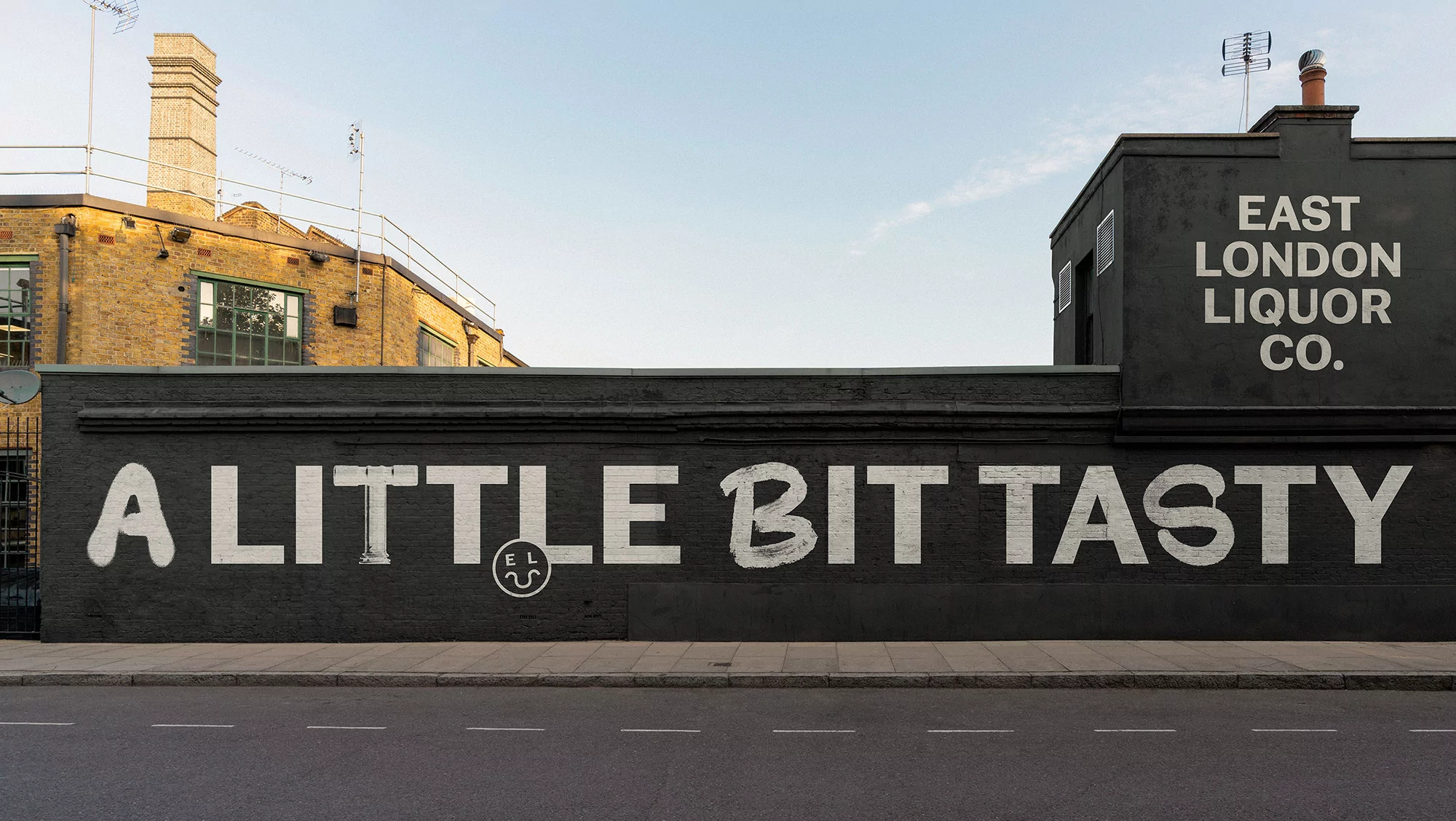 East London Liquor Co. ad on a brick building with the text 'A little bit tasty' against a blue sky and asphalt road.