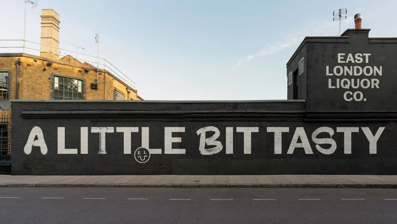 East London Liquor Co. ad on a brick building with the text 'A little bit tasty' against a blue sky and asphalt road.