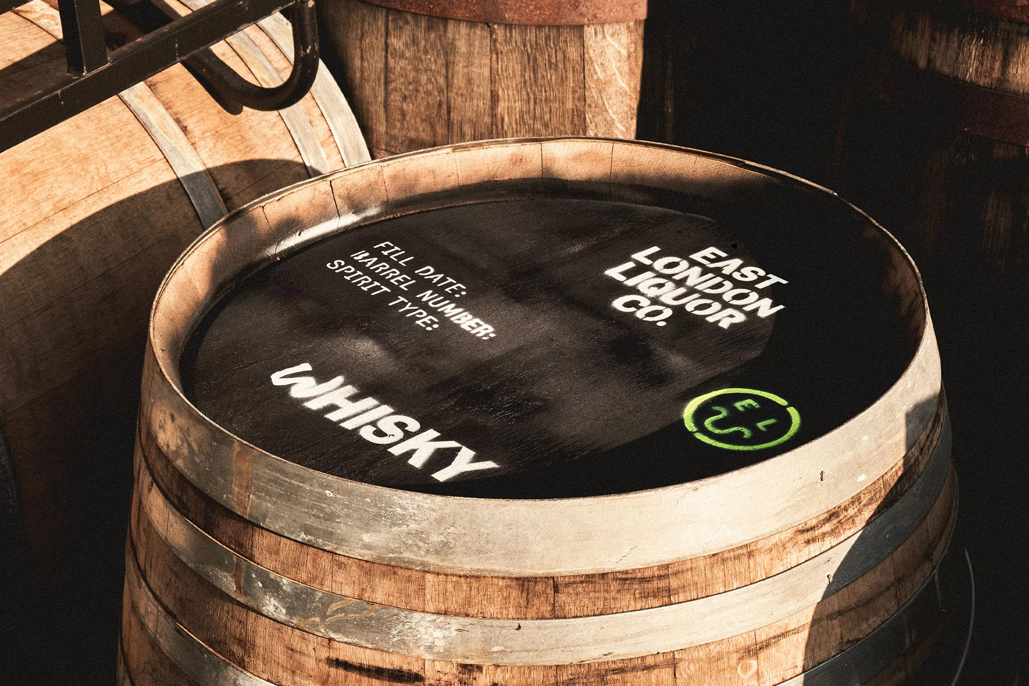 A wooden barrel with metal rings, labeled with 'East London Liquor Co.' and 'Whisky,' surrounded by other wooden barrels in soft lighting.