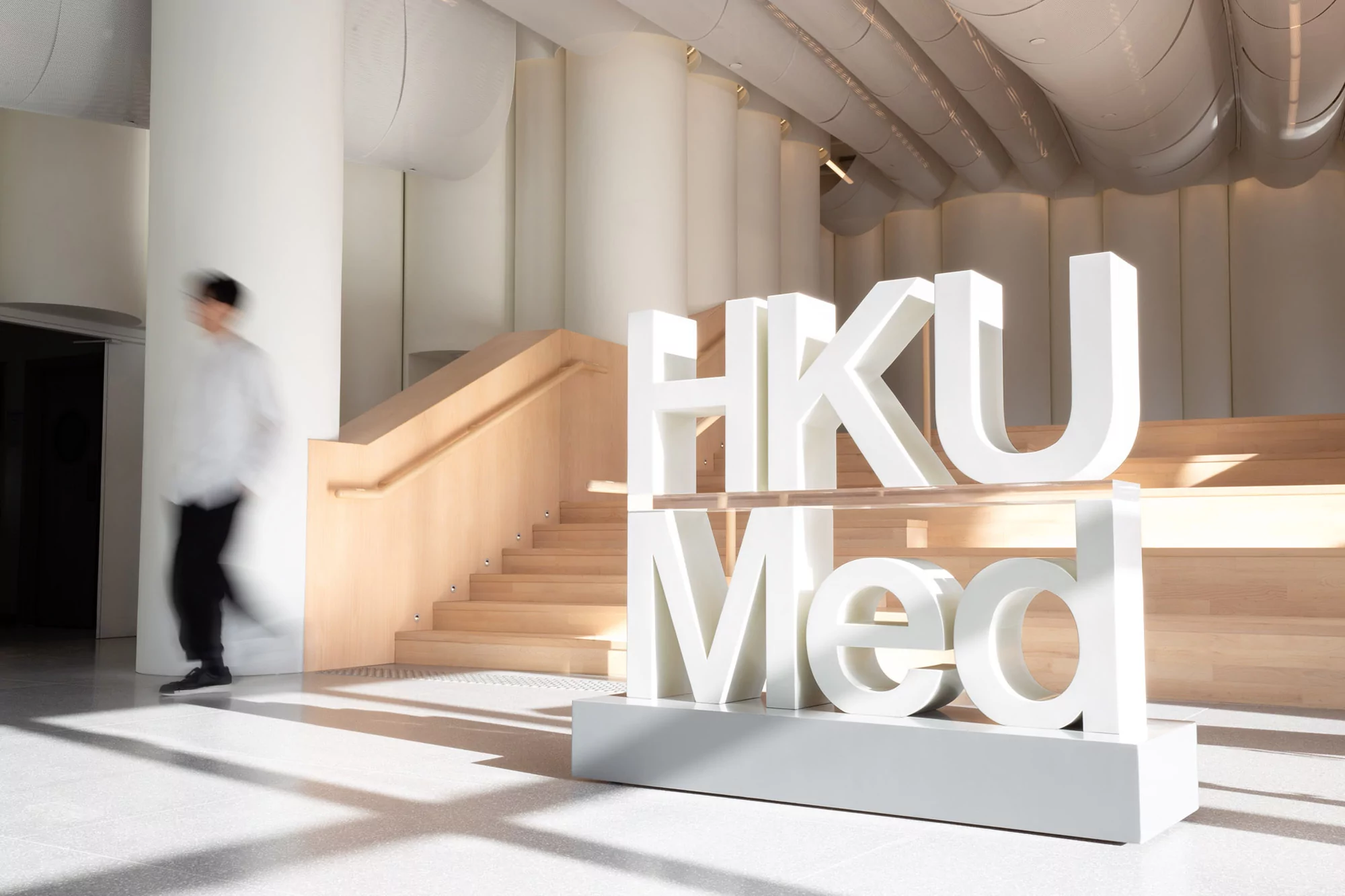 Modern interior with a blurred man passing a large HKU Med sign near wood stairs and a white column in a well-lit, spacious lobby.