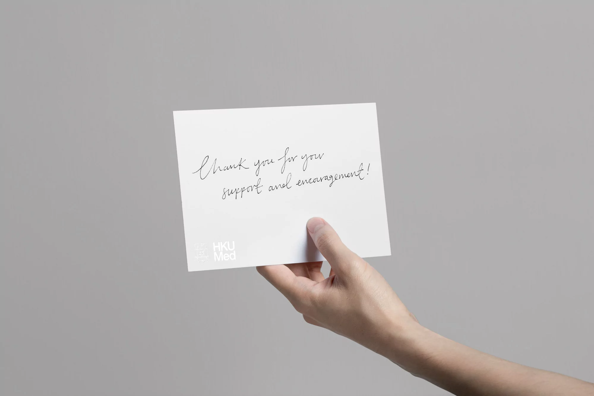 Close-up of a hand holding a white card with handwritten text, 'Thank you for your support and encouragement!', against a grey background.