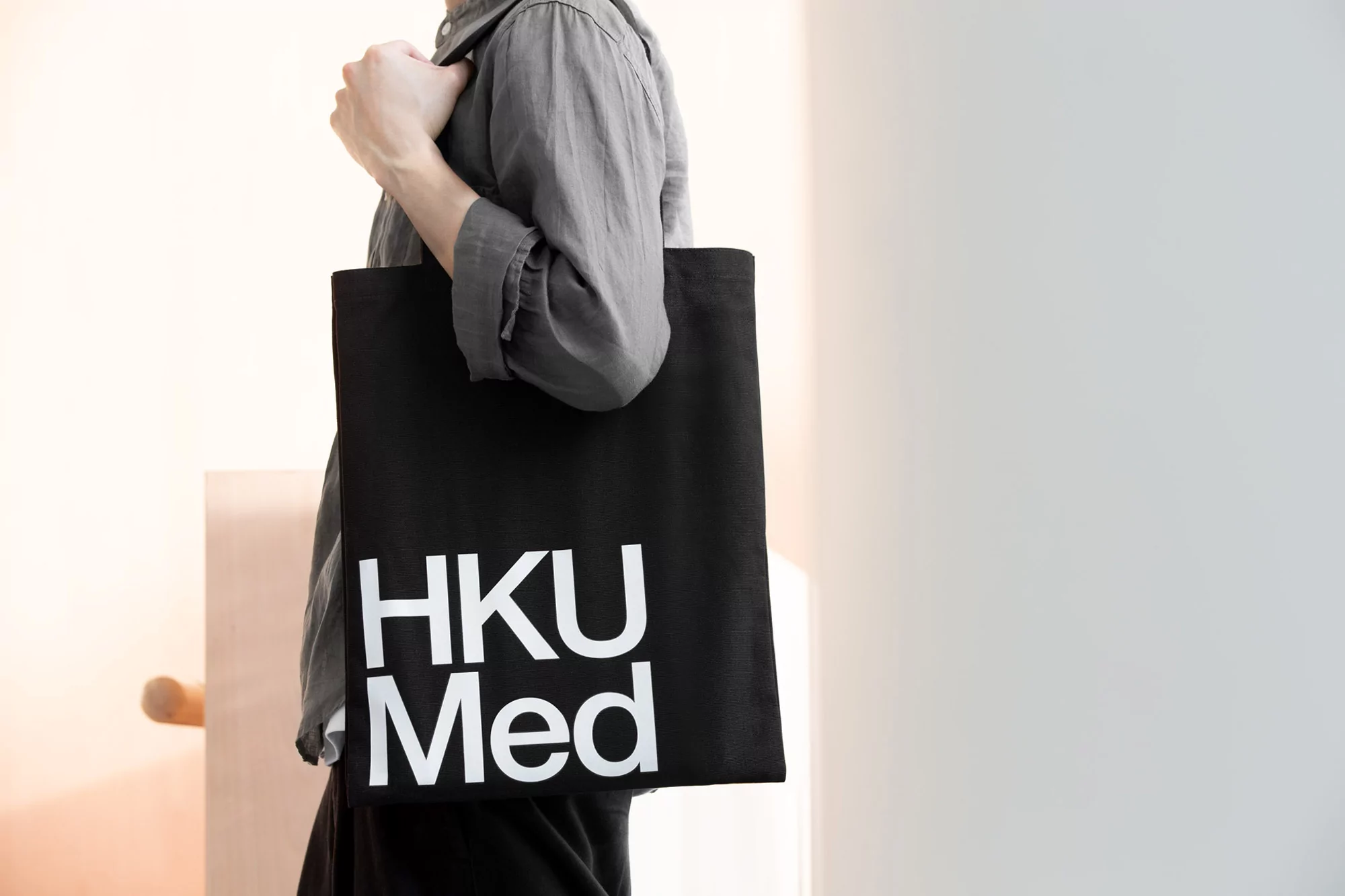 Person carries a black tote bag with 'HKU Med' printed in white, indoors. Simple wardrobe and soft, diffused lighting.