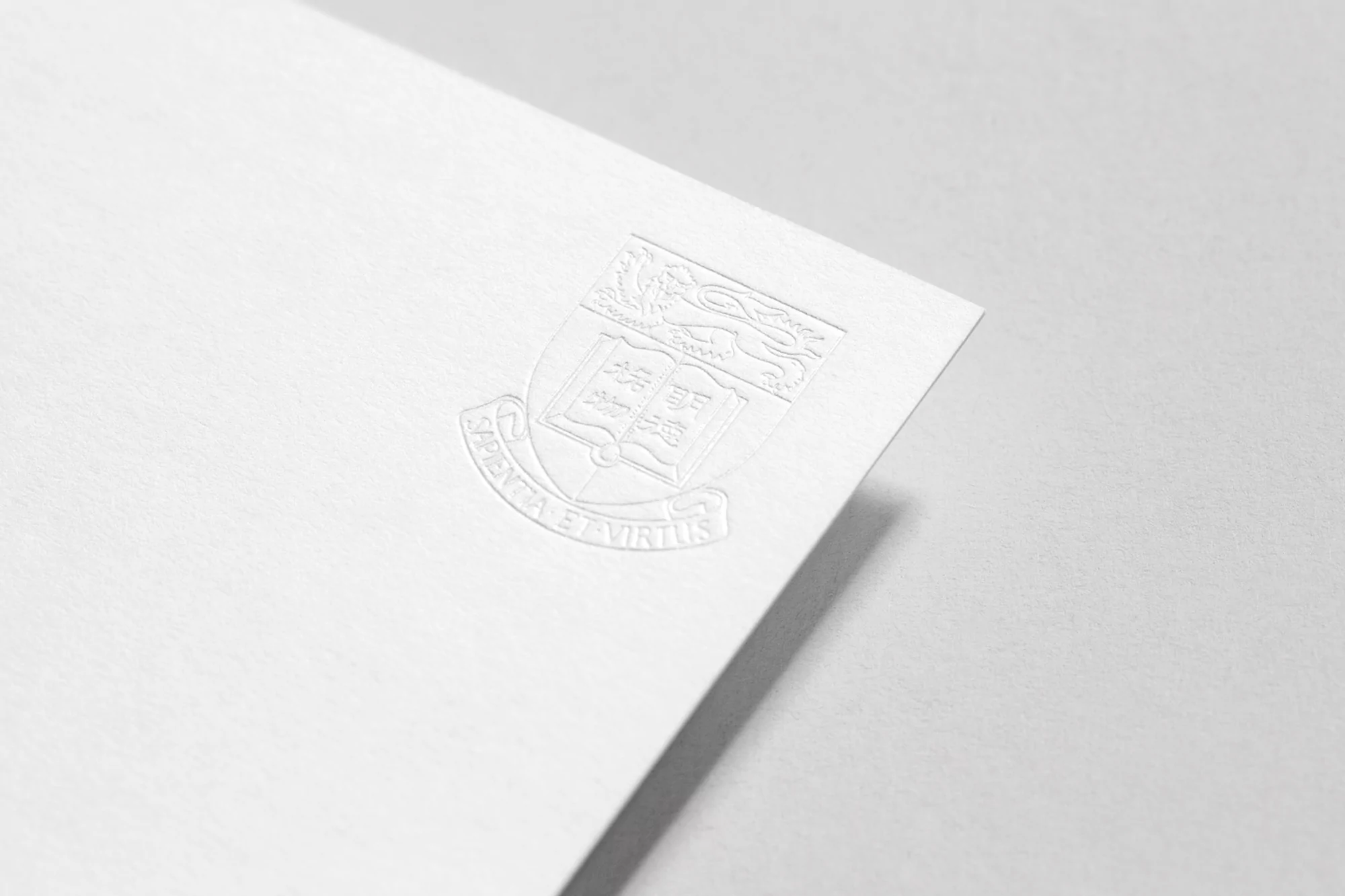 Close-up of an embossed crest on white paper, featuring a shield and Latin text, subtly textured under natural lighting.