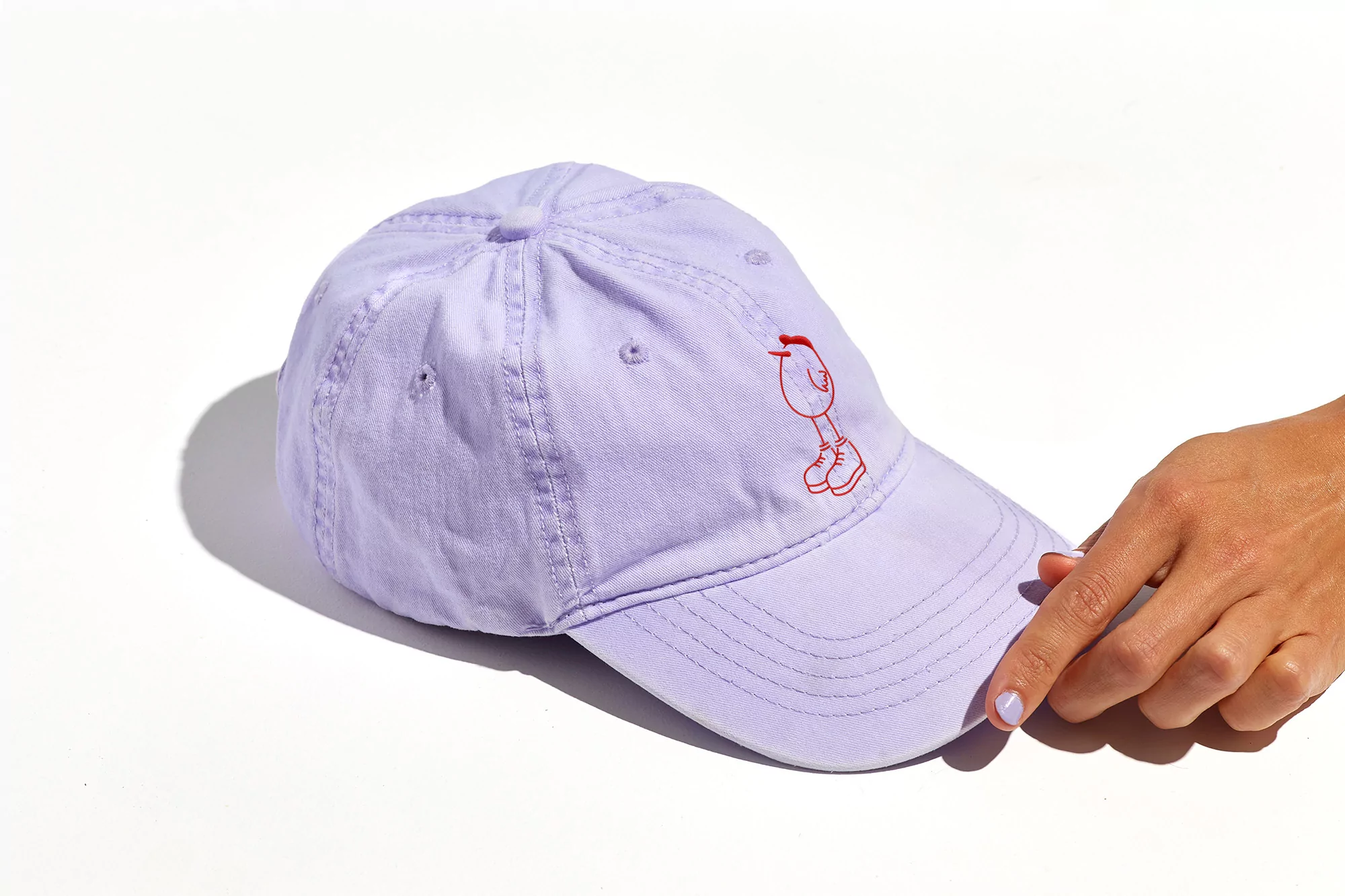Light purple baseball cap with a cartoon character on a white background, held by a hand with light skin tone, nail polish.