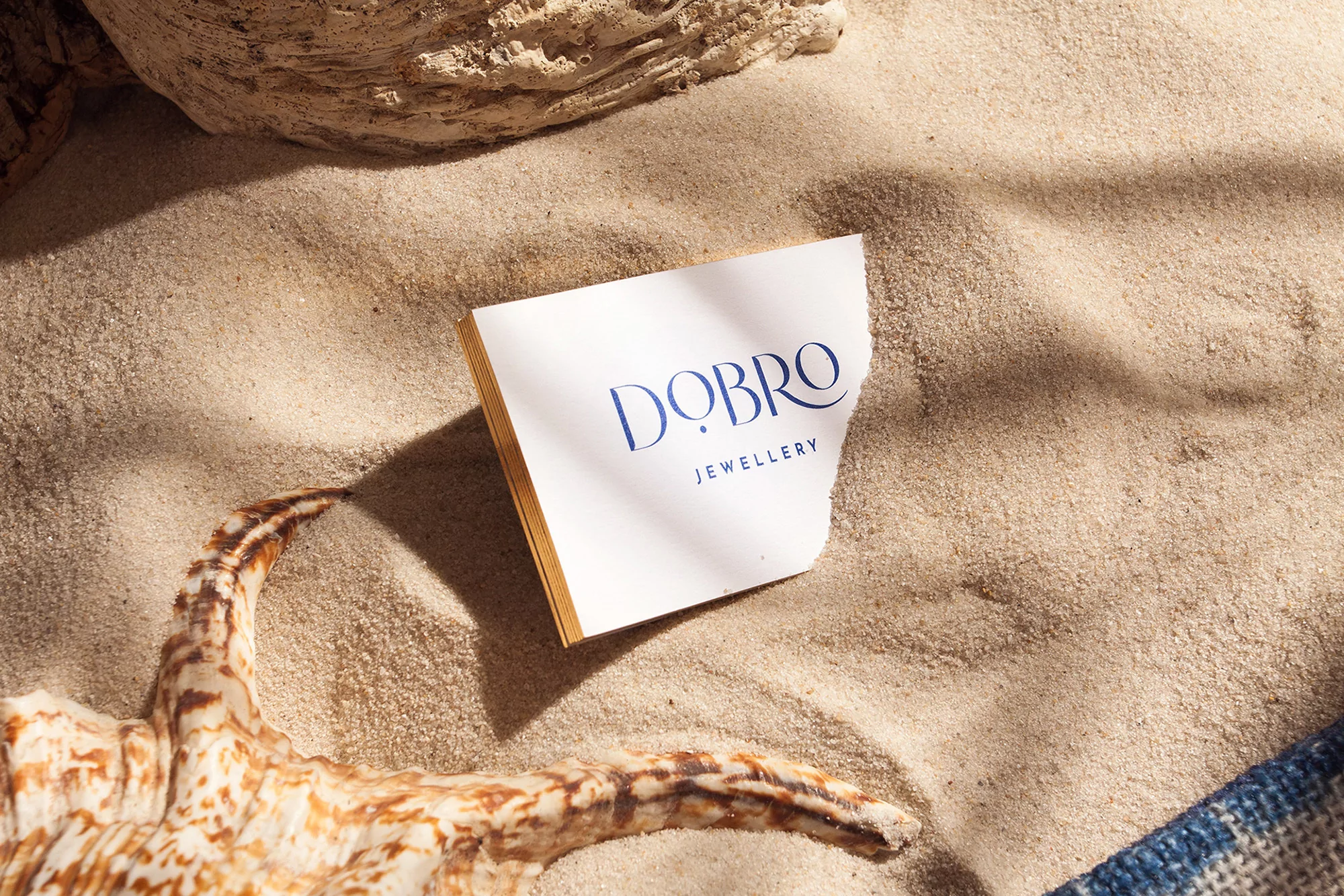 White paper card with blue Добро Jewellery text resting on sandy beach beside a seashell
