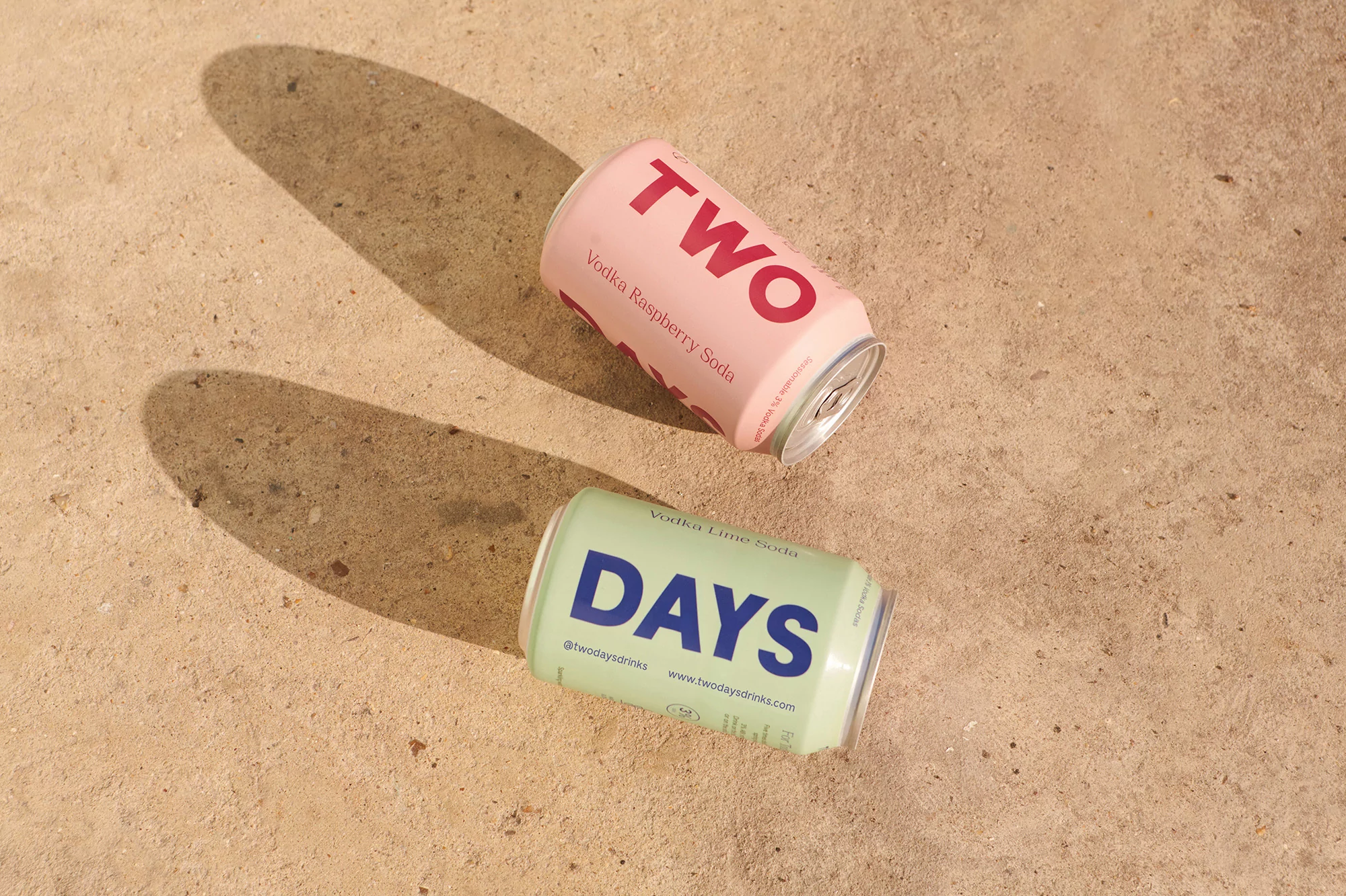 Two cans of vodka soda, one pink and one green, laying on a tan surface with shadows, creating a refreshing and inviting composition.