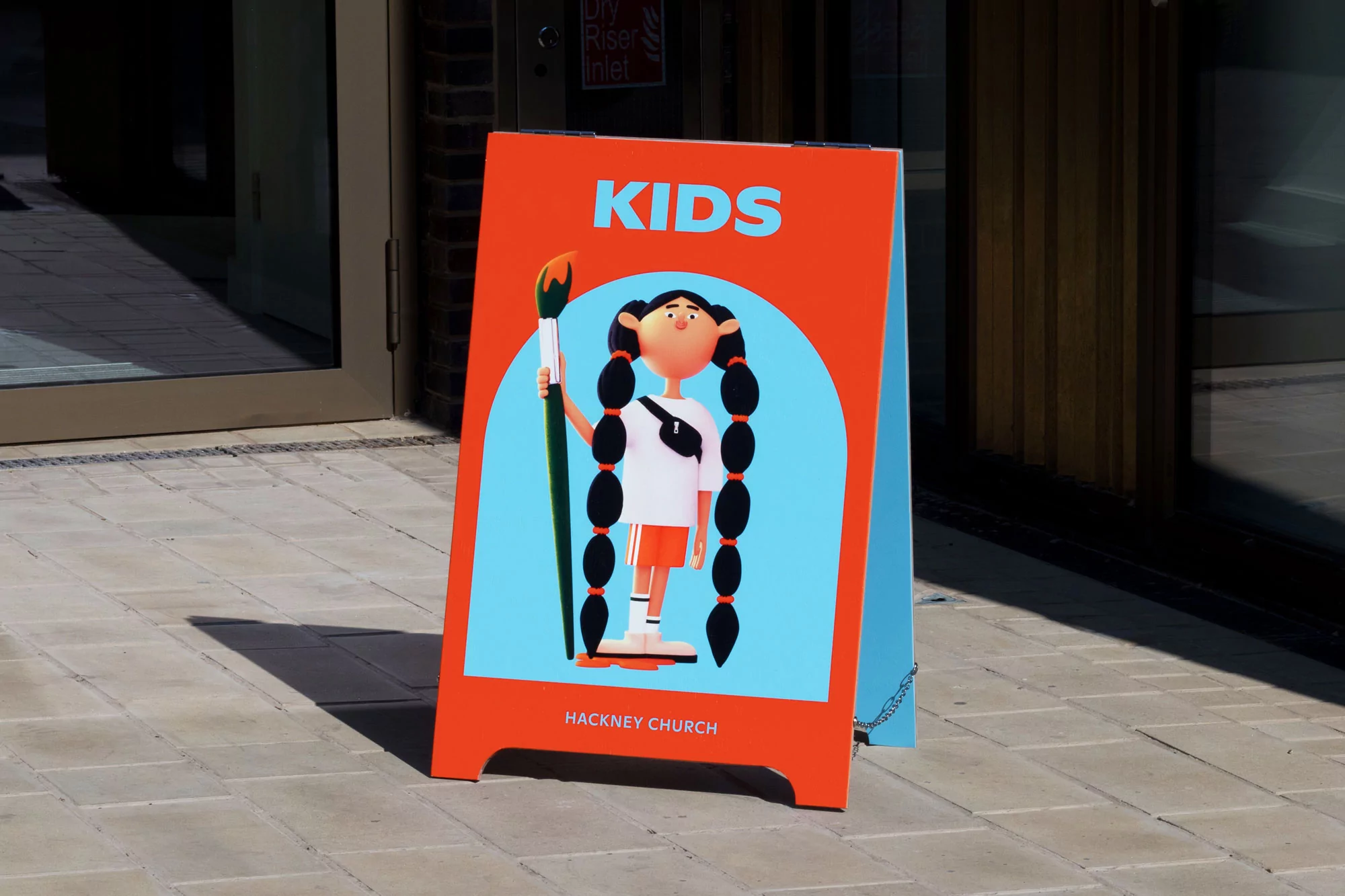 Outdoor A-frame sign for Hackney Church Kids featuring illustrated girl with braids, holding paintbrush, on a city street.