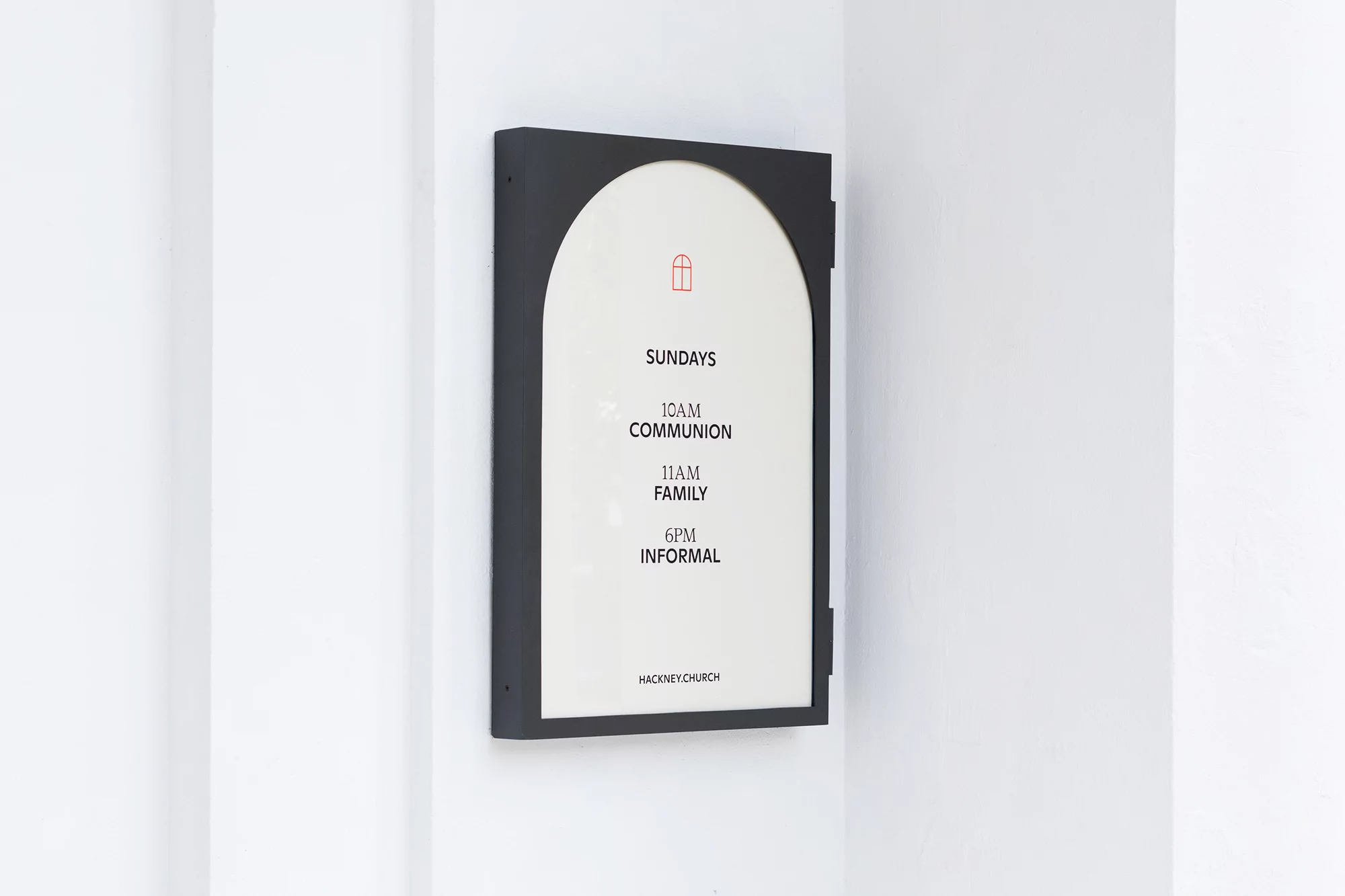 Framed sign on white wall advertises service times for Hackney Church, featuring a simple red window graphic at the top.