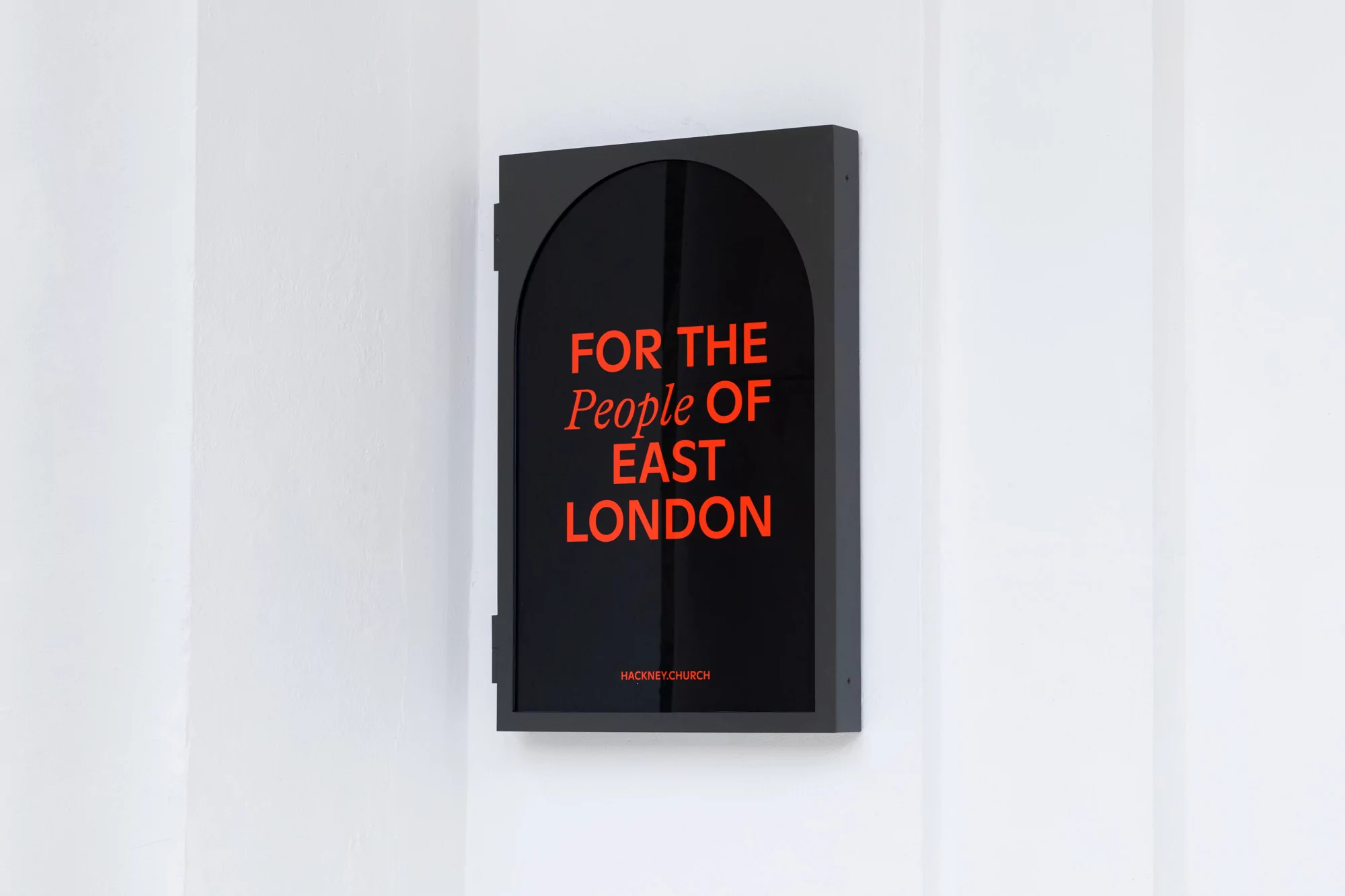 Black box-shaped sign with arched top mounted to white wall. Sign has orange text stating 'For the People of East London'.