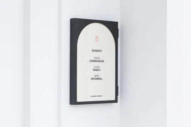 Black rectangular sign with arched window design mounted on a plain white wall displaying service times for Hackney Church.