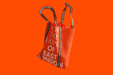 Orange tote bag featuring white text suspended against a solid orange background with branded black straps