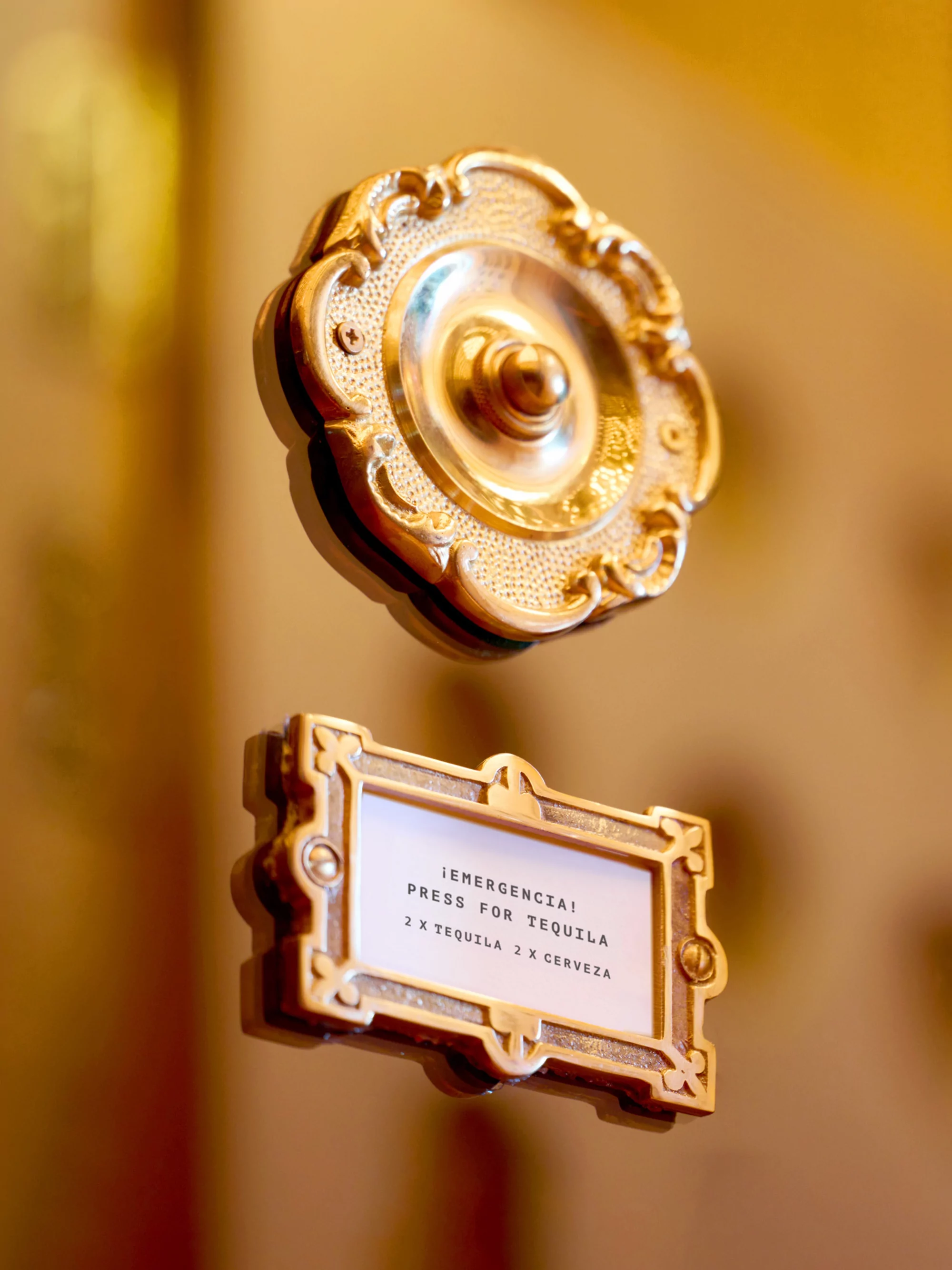 Ornate golden service bell and sign reading, '¡Emergencia! Press for Tequila, 2 x Tequila 2 x Cerveza', in a warm, neutral environment.