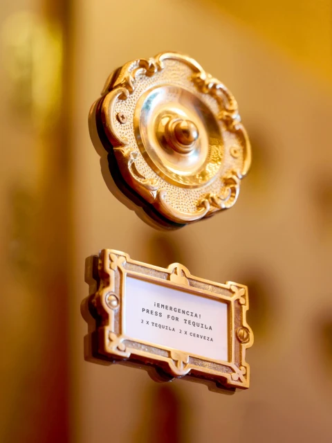 Ornate golden service bell and sign reading, '¡Emergencia! Press for Tequila, 2 x Tequila 2 x Cerveza', in a warm, neutral environment.