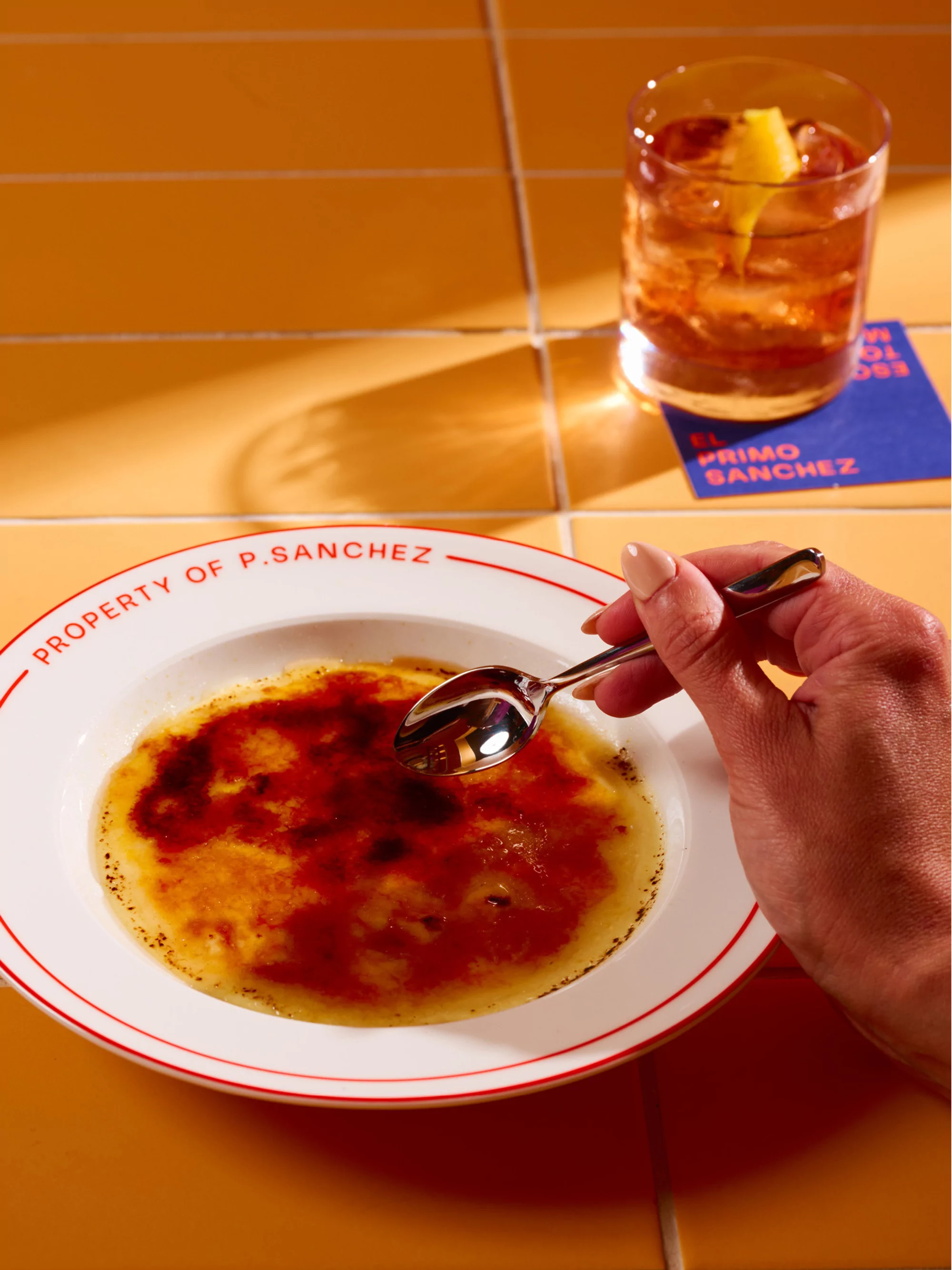 A plate of crème brûlée with a spoon, accompanied by an old-fashioned cocktail on a vibrant yellow-tiled surface, creates a tantalizing food still life.