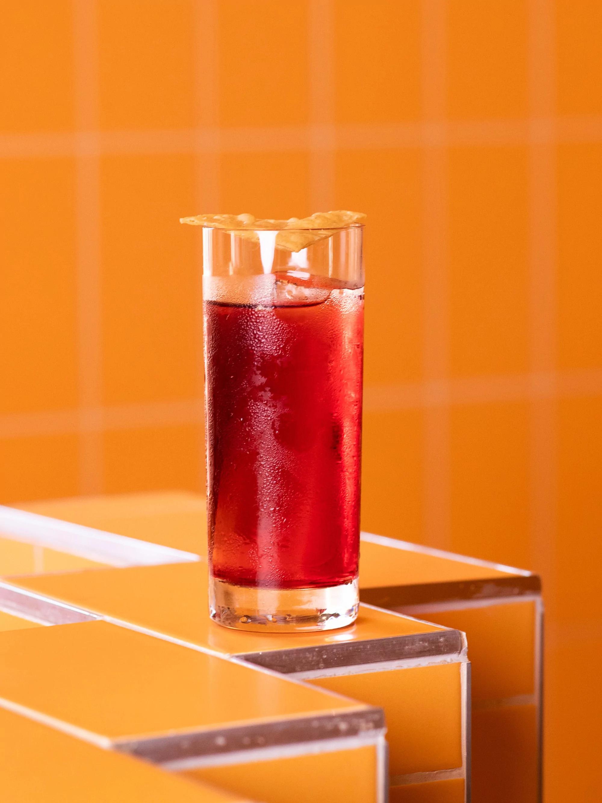 Highball glass with red cocktail and ice, garnished with a chip, on orange tile. Bright, vibrant, colorful, and visually appealing.