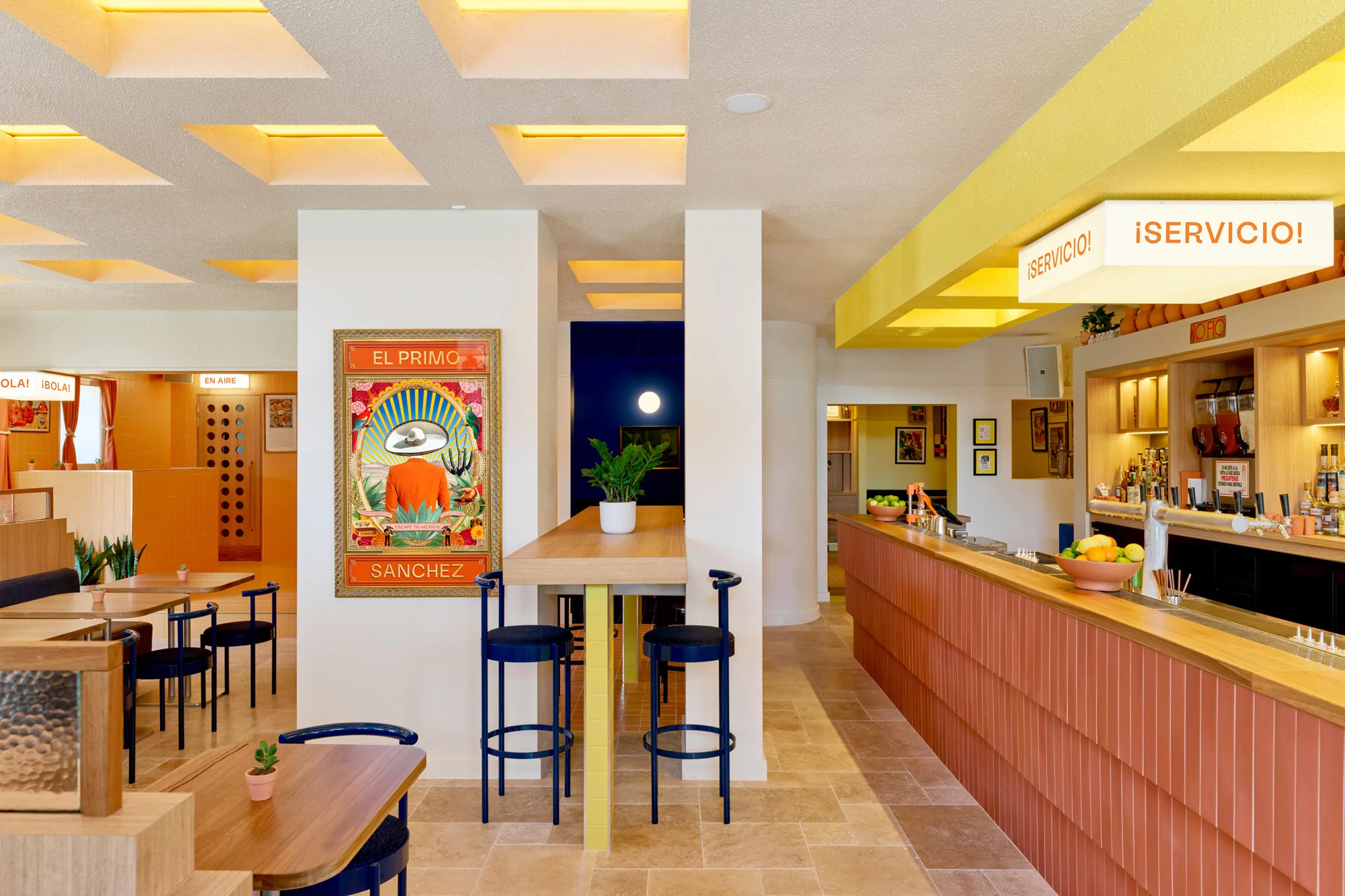 Interior view of a restaurant featuring a bar, tables, chairs, and framed artwork on the wall, illuminated by natural light.