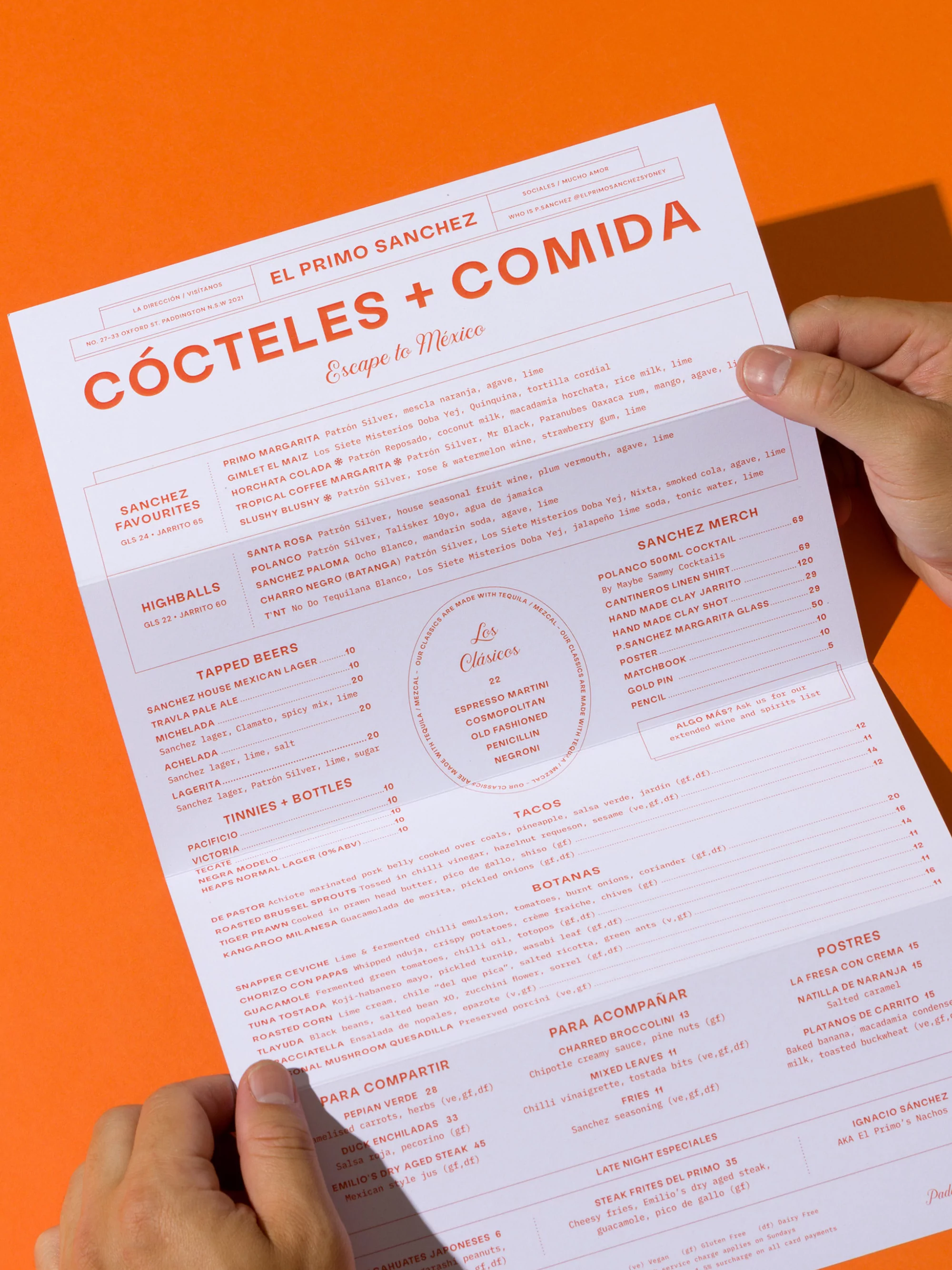 Hands holding a restaurant menu on a bright orange background. Menu features cocktails, food, and prices, with some text visible.