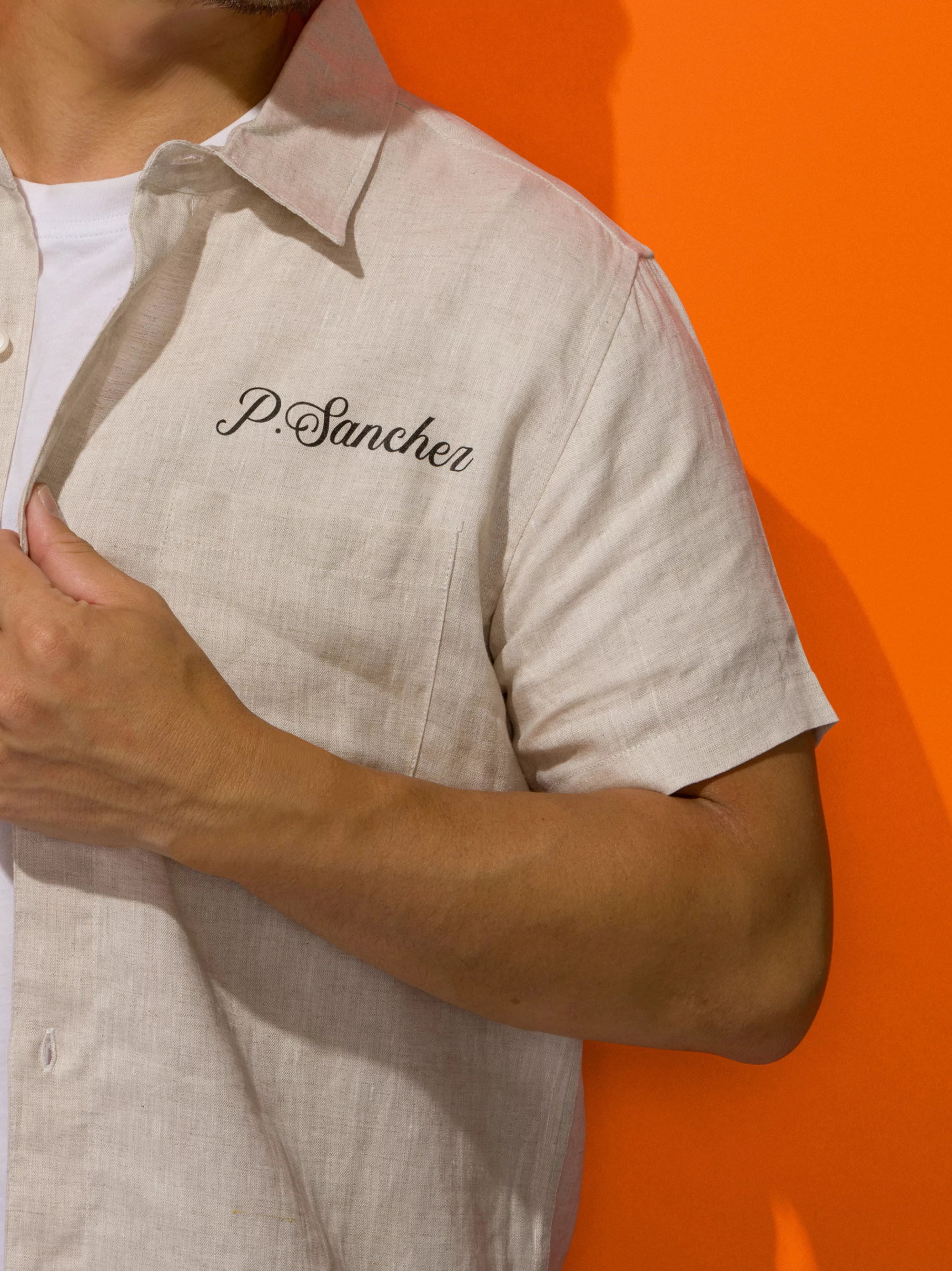 Tan linen shirt with short sleeves on an adult male with fair skin, name embroidered on the pocket against an orange background.