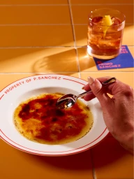 A hand holds a small spoon breaking into crème brûlée in a white bowl with red rim, alongside a cocktail on yellow tiles.