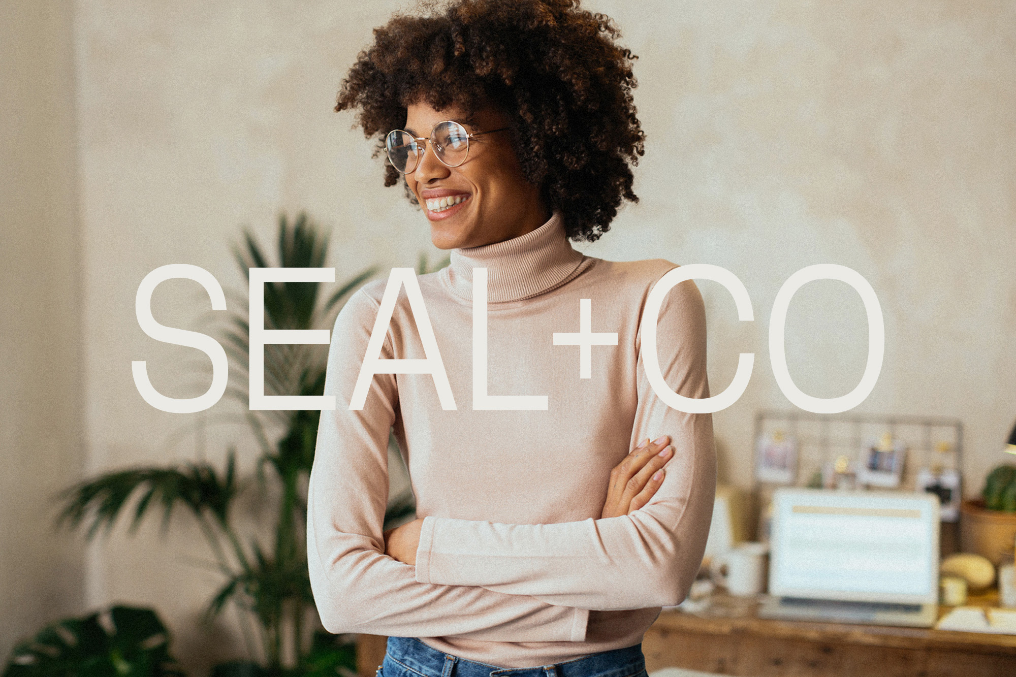 Smiling Black woman wearing glasses and a pink turtleneck, arms crossed, standing indoors near a desk with a laptop and a houseplant.