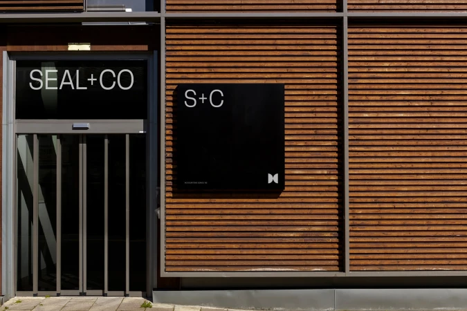 Exterior view of modern building featuring wood cladding, a black sign with 'S+C' logo, and a glass door with 'SEAL+CO' signage.