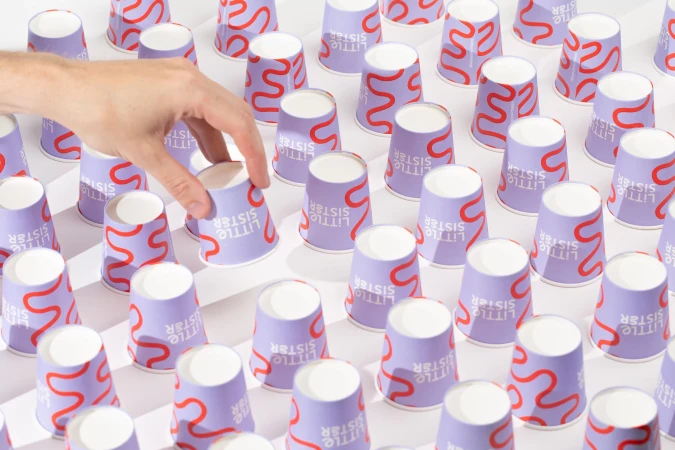 Close-up of a hand selecting a paper cup among many identically branded cups, arranged on a white surface with soft light and shadows.