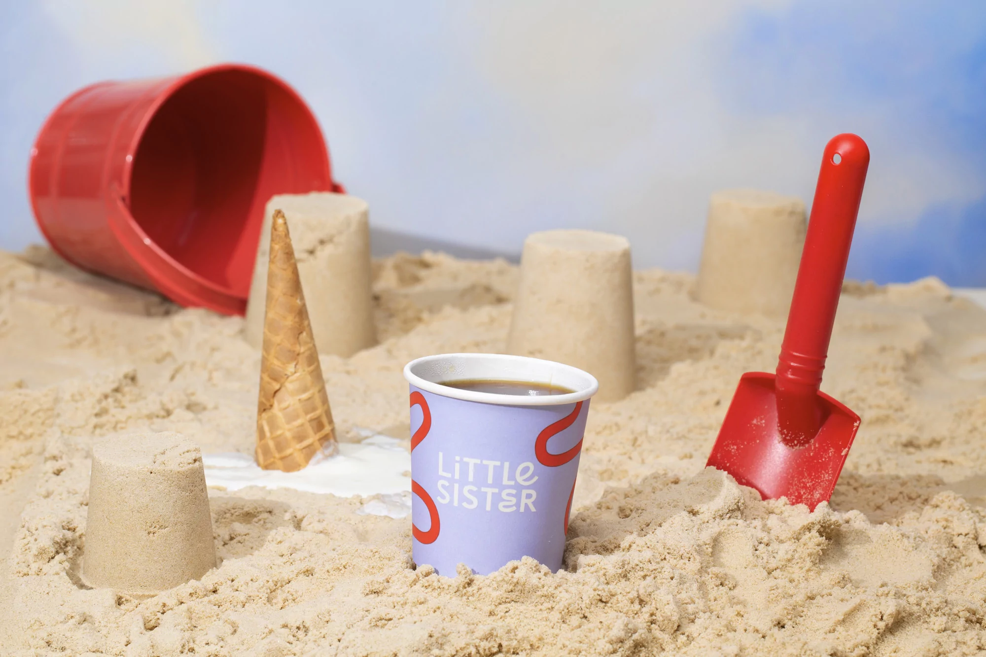 Coffee cup, sandcastle, ice cream cone, red bucket, and shovel arranged on sandy beach backdrop with blue sky in the background.