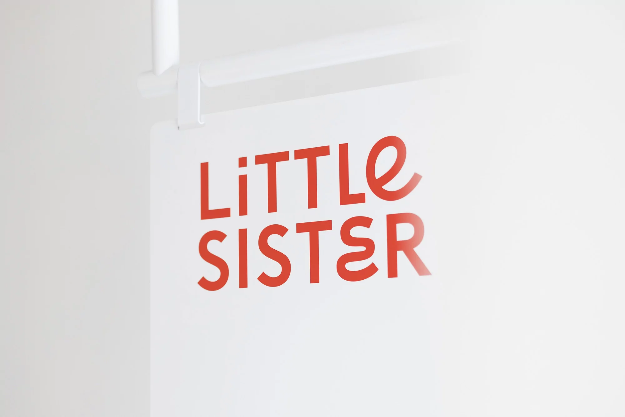 Close-up of a sign reading 'Little Sister' in red, with a white background, mounted on a white metal frame, minimalist design.