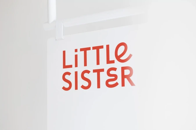 Close-up of a sign reading 'Little Sister' in red, with a white background, mounted on a white metal frame, minimalist design.