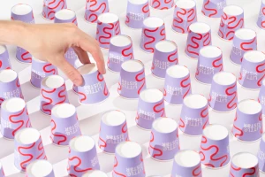 Hand picking up one purple paper cup featuring a red squiggly pattern from an organized grid arrangement on white.