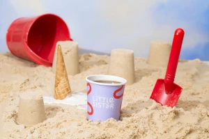 A purple paper cup with Little Sister branding sits in a sandy beach setting alongside a red bucket, toy shovel, and sandcastles.