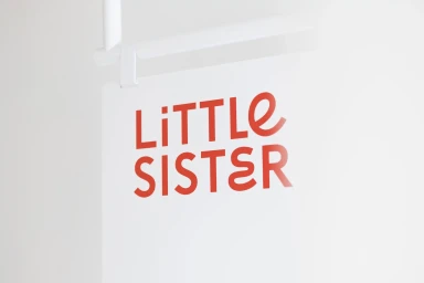 Red Little Sister brand typography displayed on a clean white hanging sign in a bright minimal studio setting