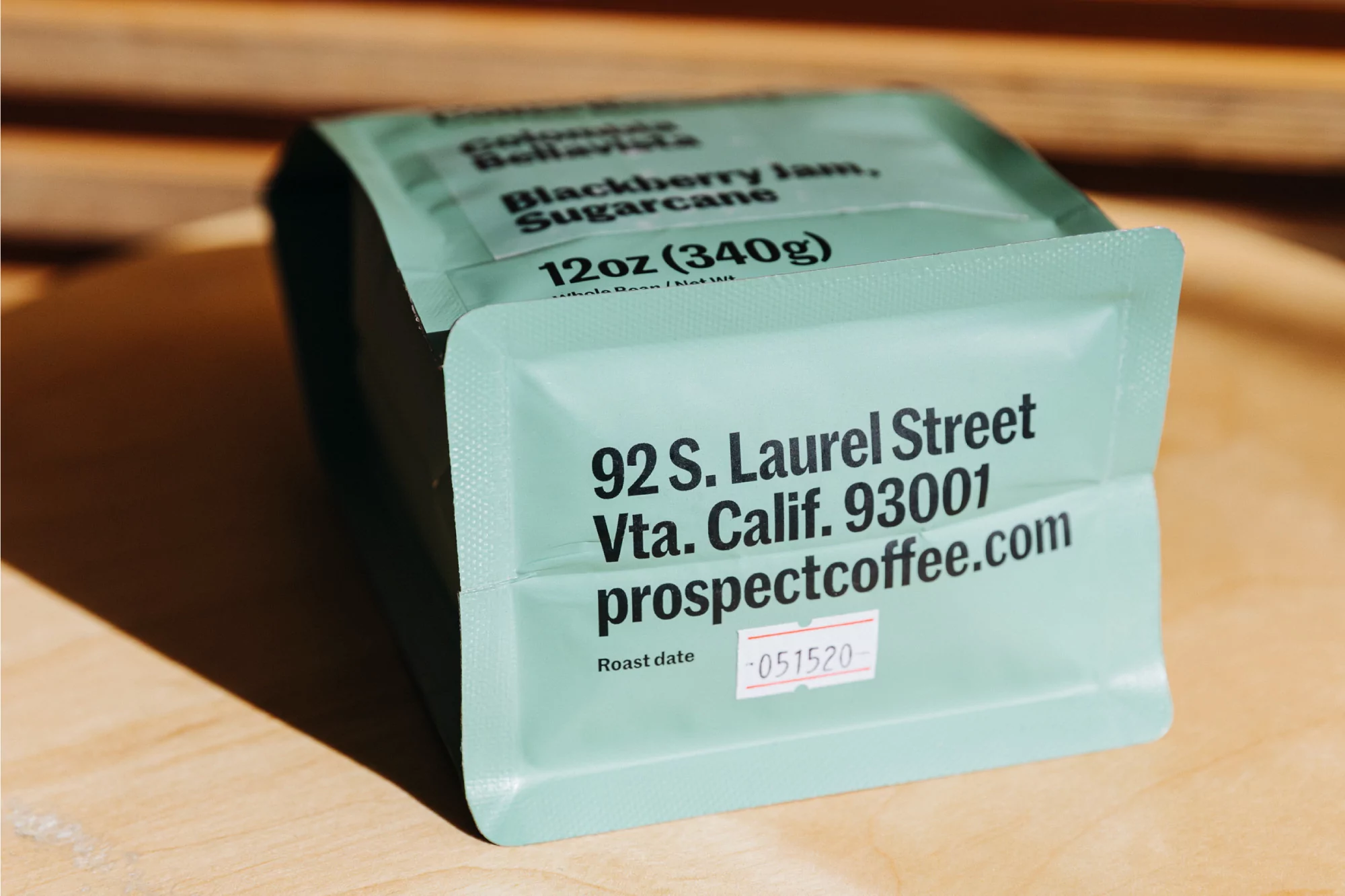 Close-up studio shot of a green bag of Prospect Coffee with details like 'Blackberry Jam, Sugarcane,' weight, address, and roast date.