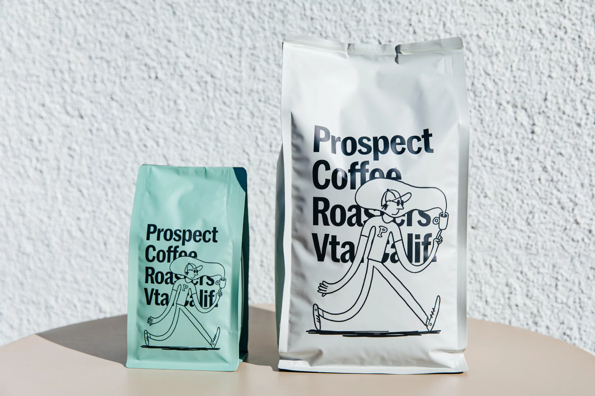 Two bags of Prospect Coffee Roasters coffee, one white and one green, stand on a table in front of a textured white wall.