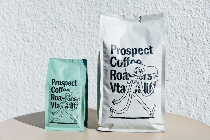 Two bags of Prospect Coffee Roasters coffee, one white and one green, stand on a table in front of a textured white wall.