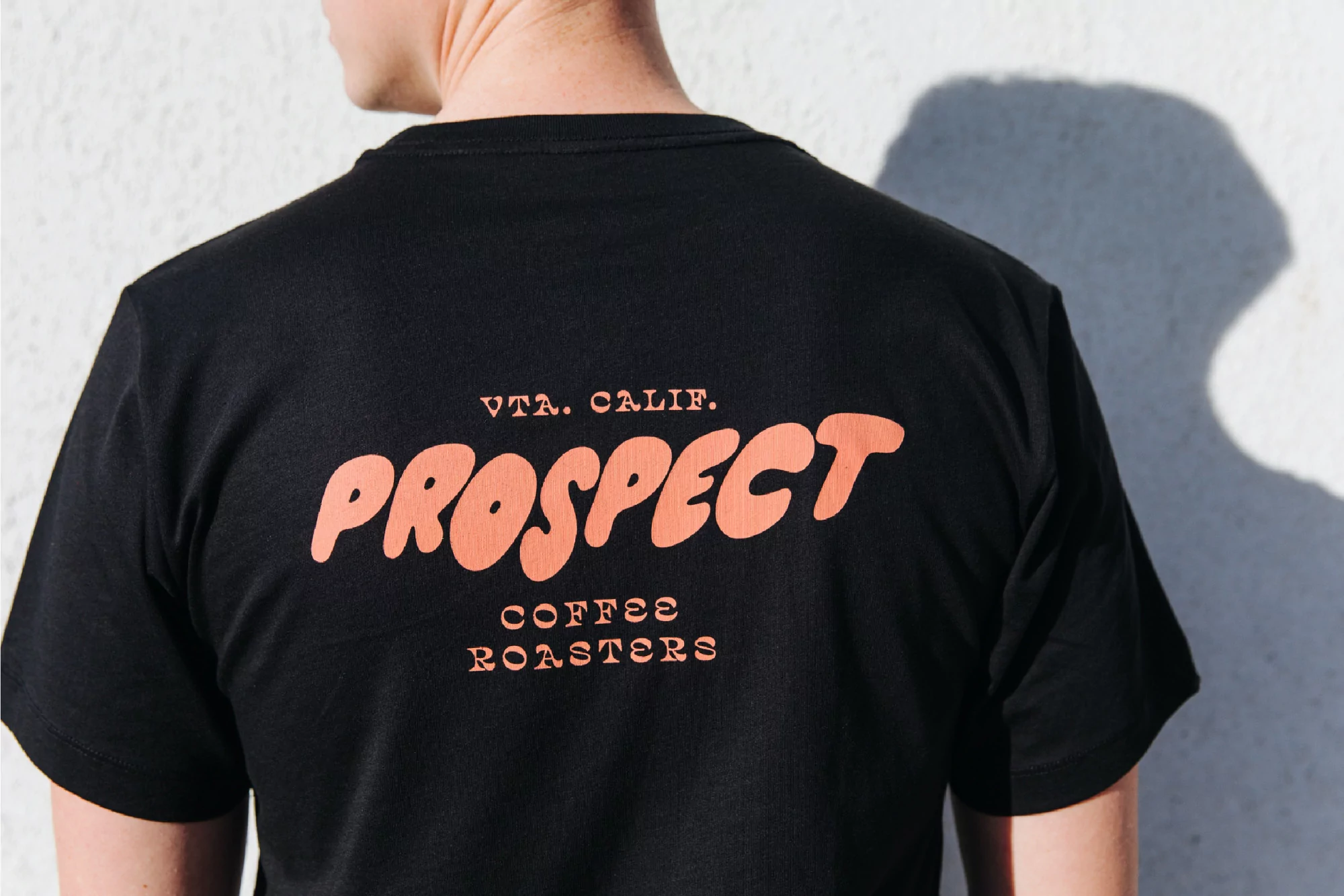 Back view of a person wearing a black T-shirt with peach-colored 'Prospect Coffee Roasters' text, standing against a light-colored wall.