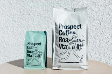 Two coffee bags with black typography and line art branding on a beige surface against a textured white wall.
