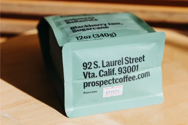Mint green coffee bag with black typography resting on a wooden surface under warm light