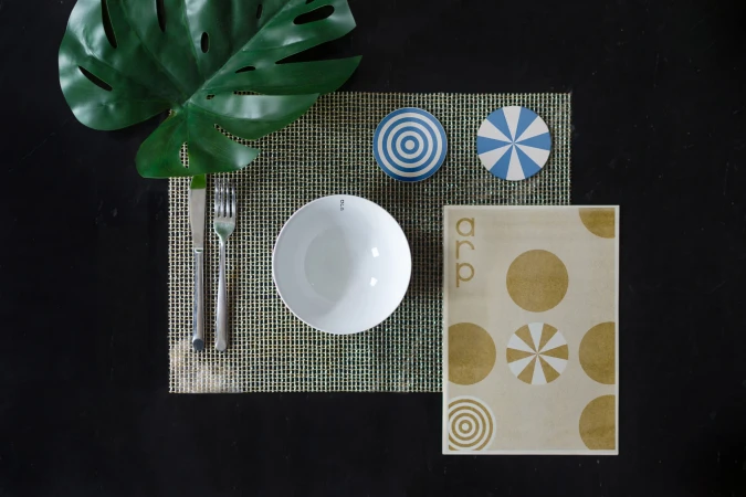 Overhead shot of a place setting with a white bowl, silverware, coasters, and a patterned napkin on a woven placemat against a black background.