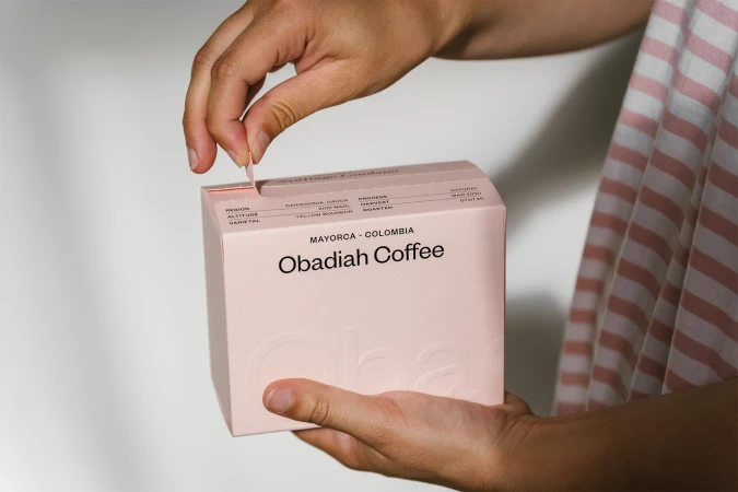 Person opening a pink box of Obadiah Coffee, close-up view. The box has text describing the region, altitude, and varietal of the coffee.