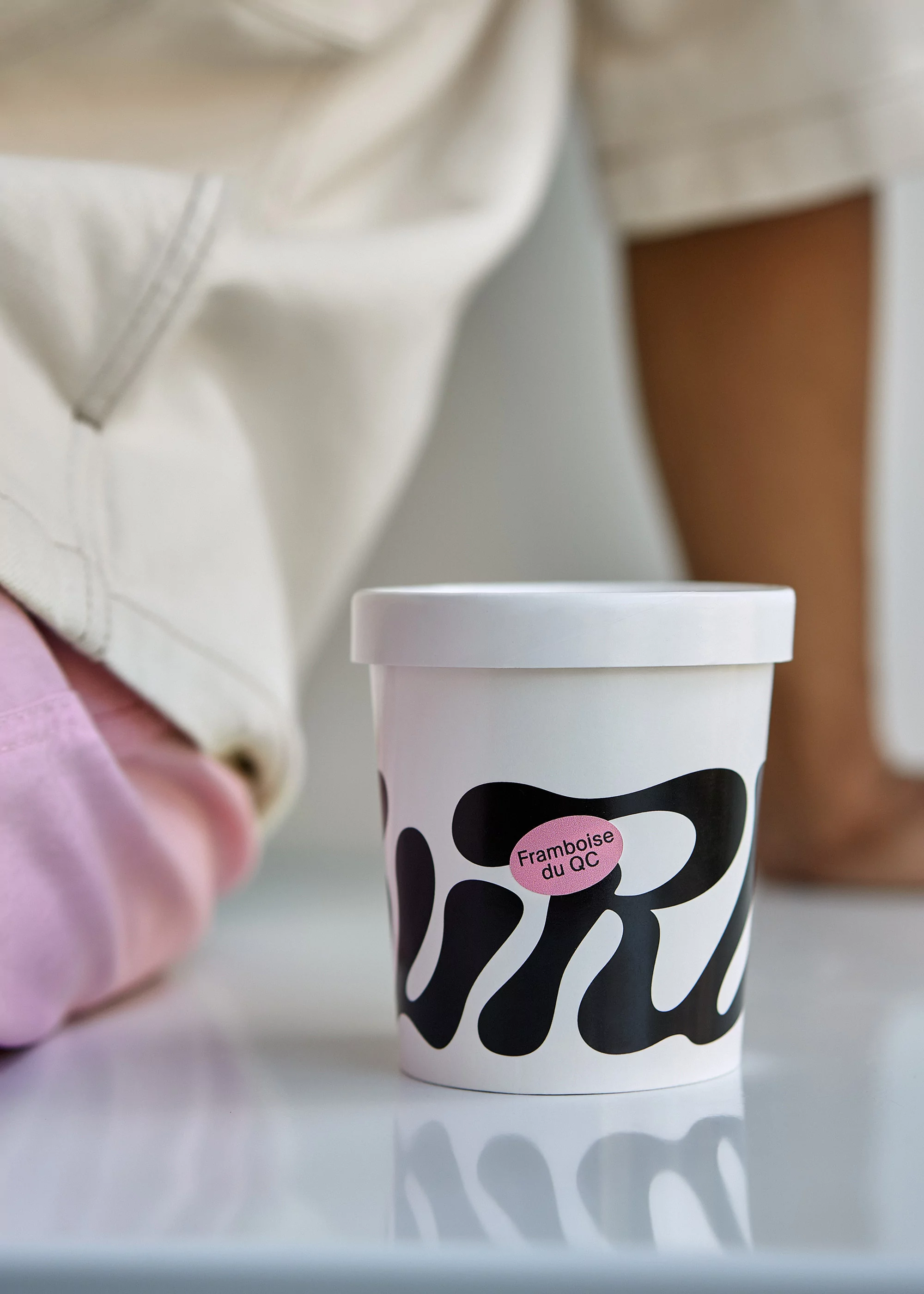 Close-up of a white container with black design and pink label, set against a neutral-toned backdrop with a blurred human figure.