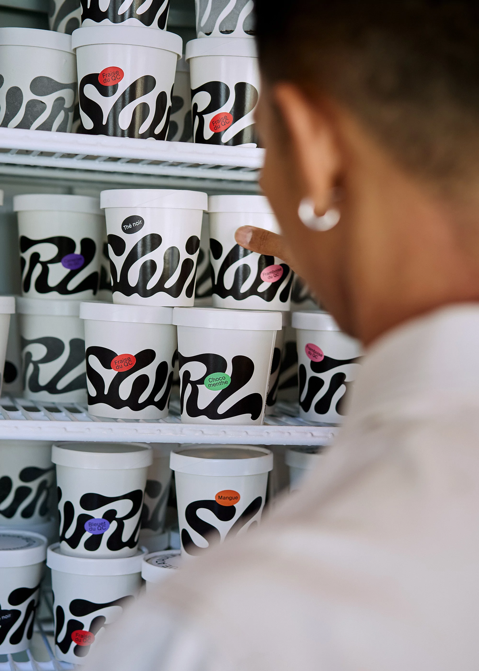 Eye-level shot of ice cream pints on refrigerated shelves. A person in a white shirt reaches out to grab one of the containers.