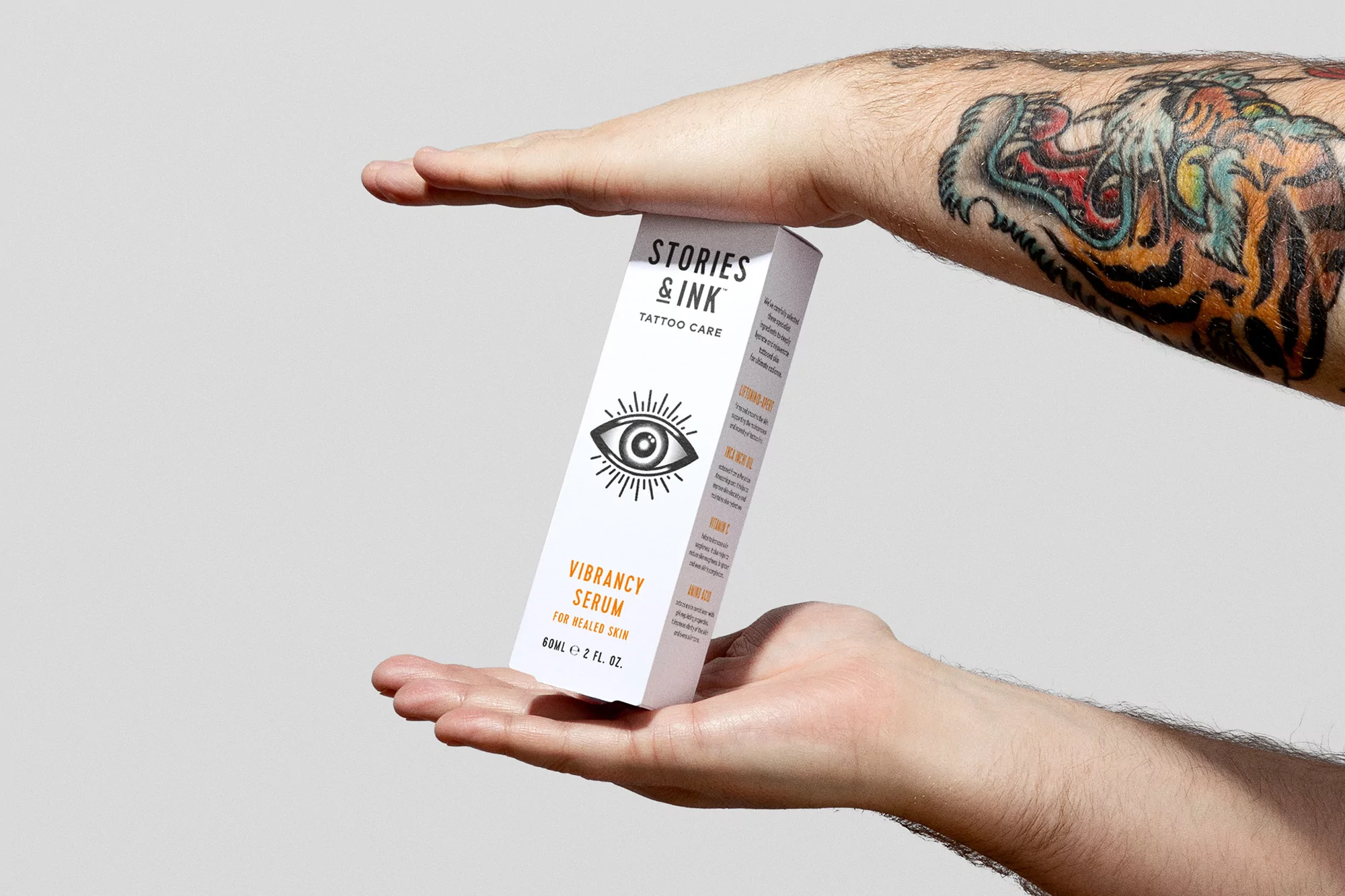 Close-up studio shot of a hand holding a white Stories & Ink Tattoo Care serum box with visible logo and text against a grey background.