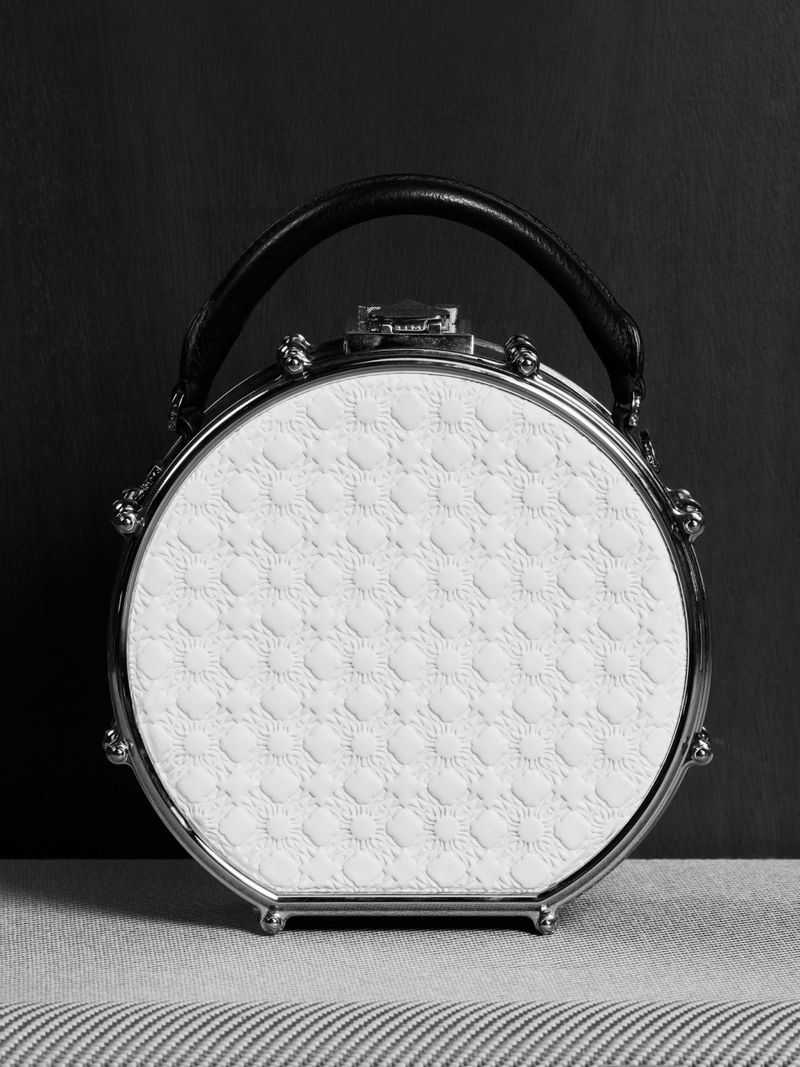 Elegant round white purse with black handle and silver hardware on a striped grey surface against a dark grey background.