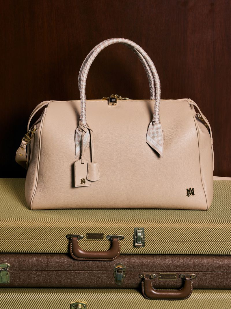Beige leather duffel bag with scarf-wrapped handles resting on stacked vintage suitcases against a dark brown background.