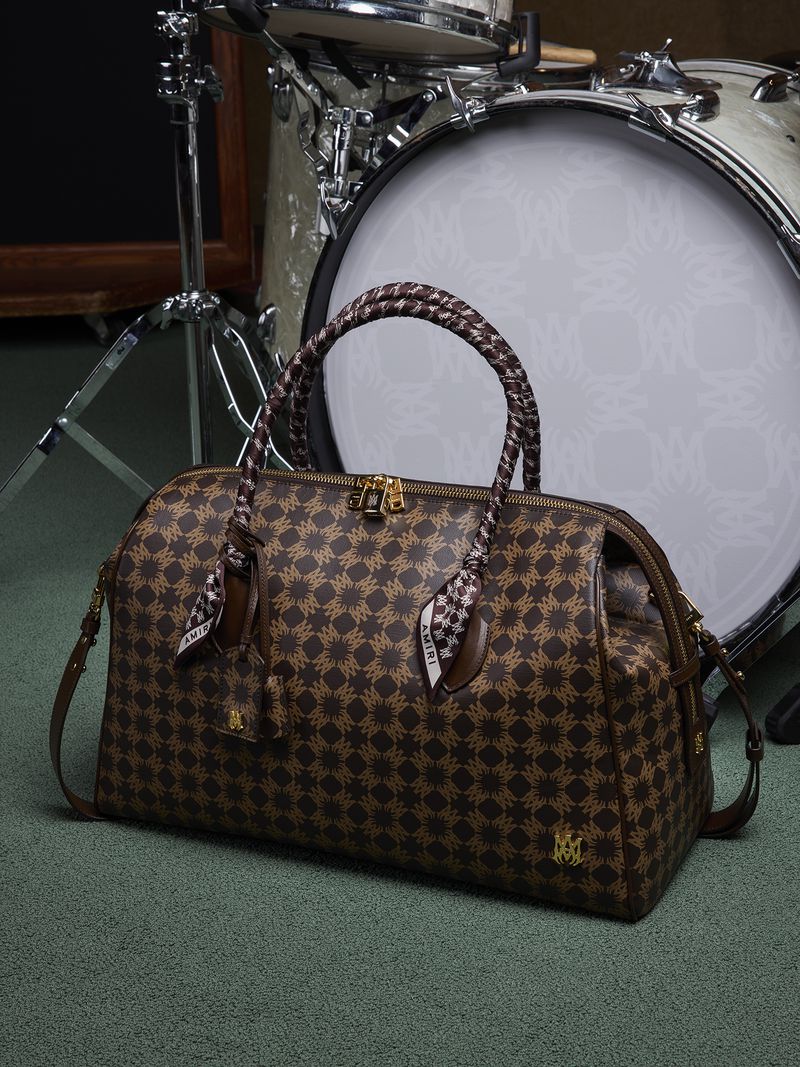Patterned barrel bag is displayed in a studio setting with a drumkit. The bag has braided handles and an optional shoulder strap.