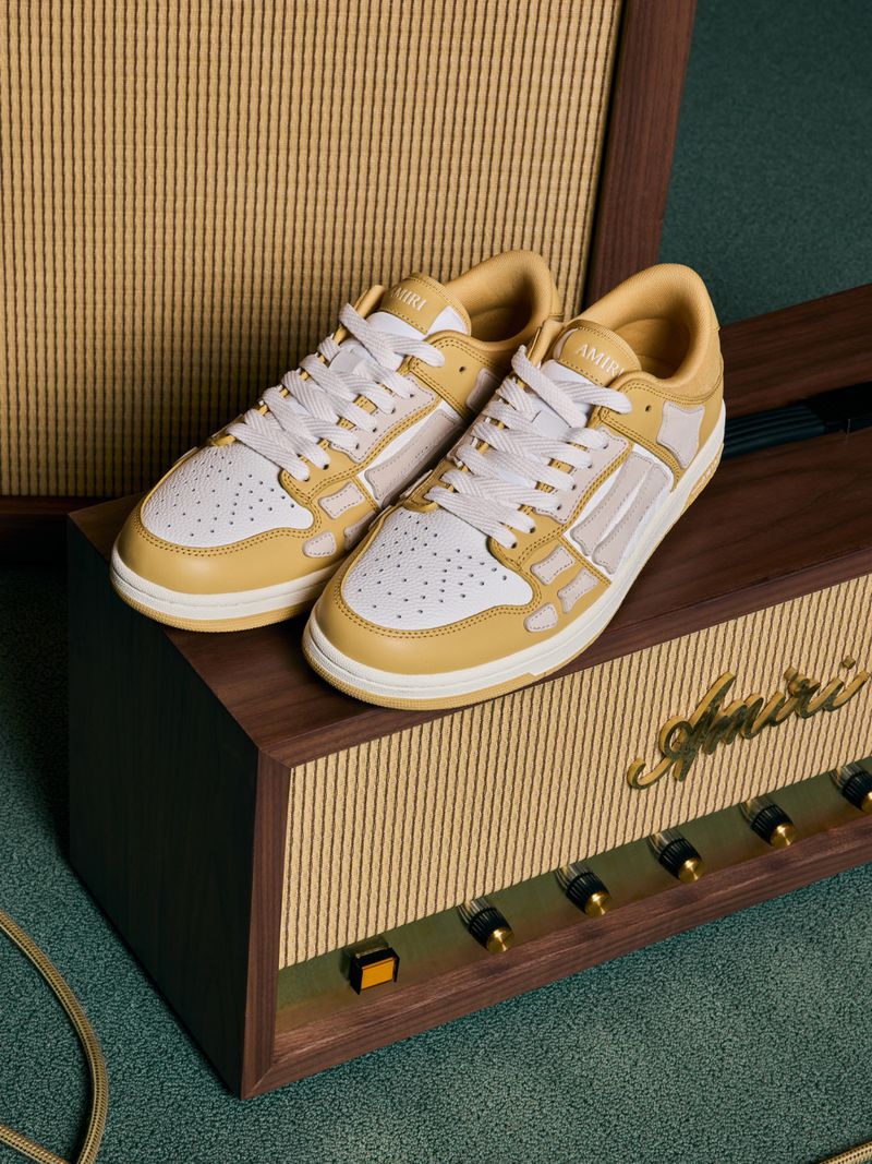 Two AMIRI bone runner sneakers displayed on an amplifier with a wooden cabinet, showcasing the brand's footwear design.