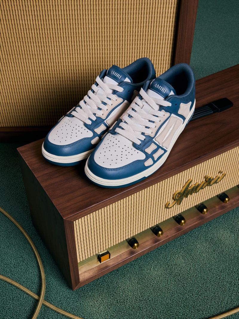 Amiri blue and white bone runner sneakers sit on top of a vintage wood and gold amplifier in a studio setting with green carpet.